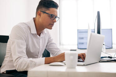 Software developer sitting at his office desk working on laptop computer. Man wearing spectacles working on laptop computer in office with a coffee cup on the table. - JLPSF15604