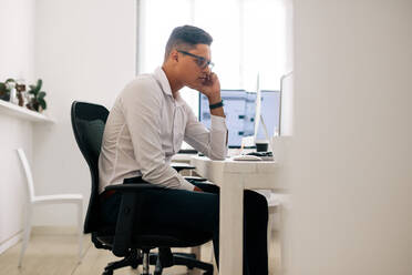 Software developer sitting at his office desk looking at the computer and thinking with hand on cheek. Man wearing spectacles working on computer in office with a coffee cup on the table. - JLPSF15605