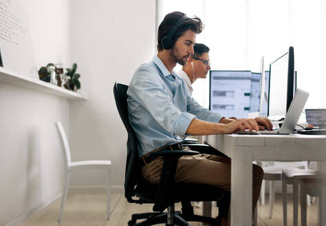 Softwareentwickler sitzen im Büro und arbeiten mit Kopfhörern an Computern. Anwendungsentwickler arbeiten an einem Laptop im Büro. - JLPSF15608
