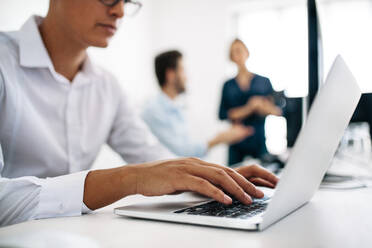 Softwareentwickler sitzen im Büro und arbeiten an Computern. Nahaufnahme eines Anwendungsentwicklers, der an einem Laptop im Büro arbeitet, mit seinen Kollegen im Hintergrund. - JLPSF15611
