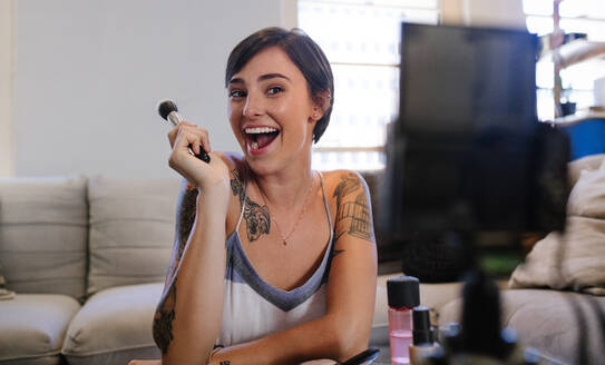 Aufgeregte junge Frau, die ihren Videoblog über neue Kosmetikprodukte aufnimmt, während sie zu Hause sitzt. Weibliche Beauty-Bloggerin zeigt, wie man ein Video-Tutorial über Make-up macht, während sie mit dem Smartphone aufnimmt - JLPSF15644