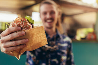 Nahaufnahme eines jungen Hipster-Mannes, der einen Burger in den Händen hält; Fokus auf die Hände, die den Burger eines Food Trucks halten. - JLPSF15668