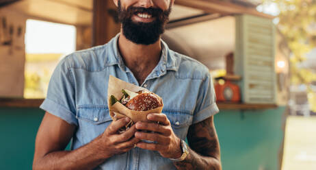 Close up of young man eating burger outdoors. Man with beard having street food. - JLPSF15669