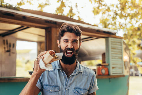 Junger Mann mit Bart isst einen Burger. Mann isst einen leckeren Burger im Freien. - JLPSF15670