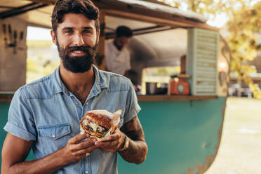 Porträt eines glücklichen jungen Mannes, der in der Nähe eines Imbisswagens mit einem Burger steht. Hipster isst einen leckeren Burger im Freien. - JLPSF15671