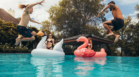 Zwei junge Männer springen mit Mädchen auf großen aufblasbaren Spielzeugen in den Pool. Gruppe junger Leute, die sich in einem Schwimmbad vergnügen. - JLPSF15674