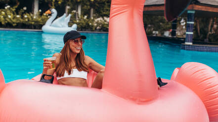 Junge Frau im Bikini sitzt entspannt auf aufblasbarem Poolspielzeug und trinkt Bier. Frau genießt auf Poolparty mit Bier. - JLPSF15678