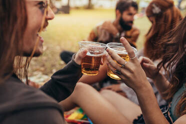 Nahaufnahme eines Paares, das mit einem Glas Bier anstößt, mit Freunden im Hintergrund. Gruppe von Leuten, die auf einem Sommerfest ein Bier trinken. - JLPSF15681