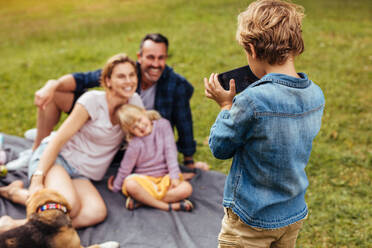 Kleiner Junge fotografiert seine fröhliche Familie auf einer Decke sitzend im Park. Sohn fotografiert Familie beim Picknick im Park. - JLPSF15728