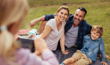 Cheerful family sitting on at park and looking at little girl taking pictures using mobile phone. Daughter photographing family during picnic at park. - JLPSF15729