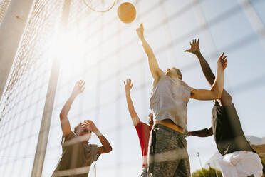 Four men playing basketball in a basketball court on a sunny day. Men jumping high to reach for the ball near the basket. - JLPSF15745