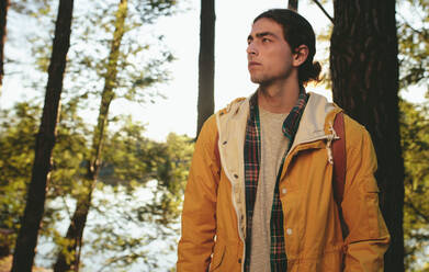 Mann auf einem Abenteuertrip, der in einem Wald steht und in die Ferne schaut. Porträt eines jungen Mannes mit Jacke, der die Wildnis erkundet. - JLPSF15746