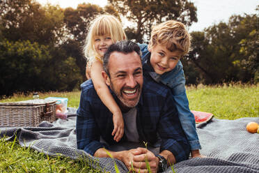 Happy man with kids on a picnic lying down in park beside a picnic basket. Children lying on the back of their father. - JLPSF15750