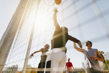 Vier Männer spielen Basketball auf einem Basketballplatz an einem sonnigen Tag. Ein Mann springt hoch, um den Ball in den Korb zu werfen. - JLPSF15760