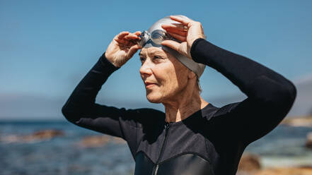 Nahaufnahme einer Frau in Badekleidung, die am Meer steht. Eine Schwimmerin steht am Strand und hält ihre Schwimmbrille auf dem Kopf. - JLPSF15766