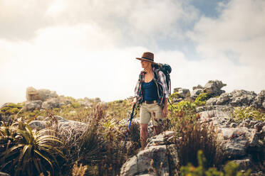 Frau geht auf einem felsigen Hügel und hält einen Trekkingstock. Frau mit Hut und Rucksack auf einer Trekkingtour. - JLPSF15771