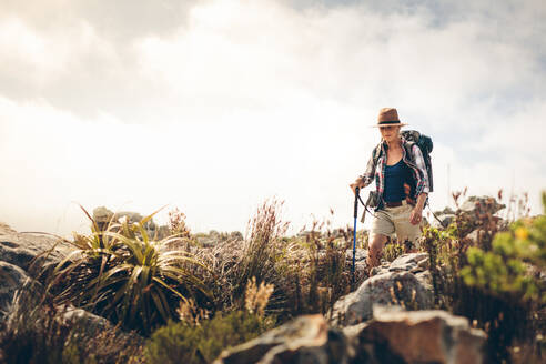 Frau, die mit Hut und Rucksack einen Hügel hinuntergeht. Frau auf einem Wanderabenteuer, die auf einem felsigen Pfad wandert und einen Trekkingstock hält. - JLPSF15772