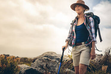 Ältere Frau mit Hut und Rucksack auf einer Wanderung in der Natur mit Wanderstock. Abenteuerlustige Frau auf einem Wanderweg an einem bewölkten Tag. - JLPSF15773