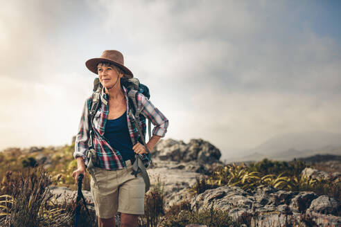 Ältere Frau wandert einen Hügel hinunter und hält einen Trekkingstock. Lächelnde Frau, die auf einem felsigen Hügel auf dem Lande wandert und einen Rucksack trägt. - JLPSF15775