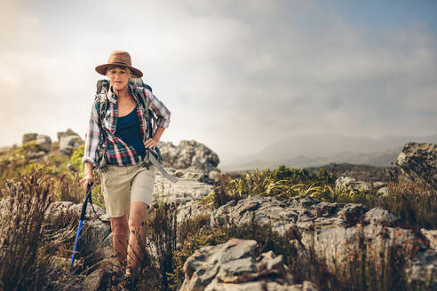 Frau mit Rucksack, die auf einem felsigen Hügel wandert. Ältere Frau, die auf einem Hügel wandert und einen Wanderstock hält. - JLPSF15776