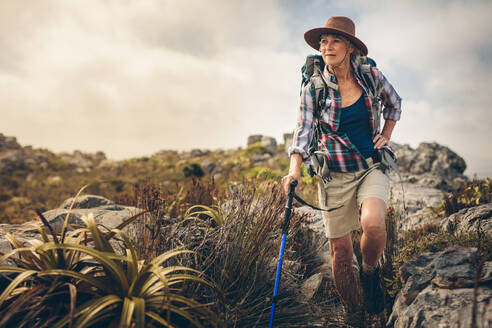 Ältere Frau, die während ihrer Wanderung durch Büsche und Felsen läuft. Wanderin, die auf einem felsigen Hügel läuft, einen Rucksack trägt und einen Wanderstock hält. - JLPSF15777