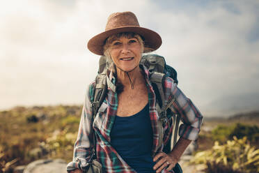 Nahaufnahme einer lächelnden Frau, die während eines Urlaubs im Freien steht. Ältere Frau mit Rucksack und Hut, die während einer Trekkingtour auf einem Hügel steht. - JLPSF15778