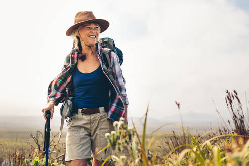 Lächelnde Frau beim Wandern auf dem Lande mit Rucksack. Ältere Frau, die auf einem Hügel wandert und einen Trekkingstock hält. - JLPSF15780