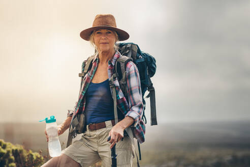 Porträt einer älteren Frau auf einem Wanderweg, die auf einem Hügel stehend Wasser trinkt. Nahaufnahme einer älteren Wanderin, die ihre Wanderausrüstung trägt und auf einem Hügel stehend eine Wasserflasche hält. - JLPSF15789