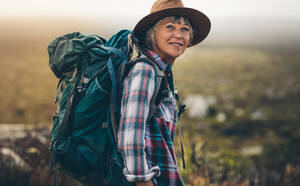 Nahaufnahme einer älteren Frau mit Hut und Rucksack im Urlaub. Seitenansicht einer Wanderin, die auf einer Anhöhe steht, während sie eine Trekkingtour macht. - JLPSF15792