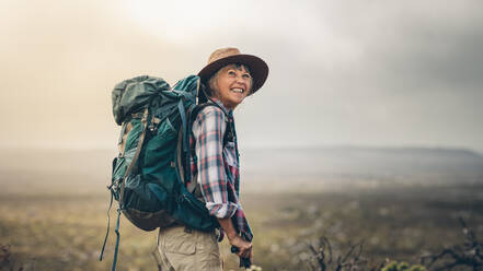 Seitenansicht einer älteren Frau mit Rucksack auf einer Anhöhe während einer Wanderung. Lächelnde ältere Frau, die während einer Wanderkampagne begeistert aussieht. - JLPSF15793