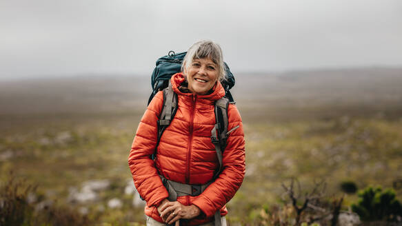 Porträt einer lächelnden Wanderin, die an einem Wintertag auf einem Hügel steht. Ältere Frau mit Jacke und Rucksack beim Wandern auf einem Hügel. - JLPSF15794