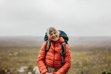 Nahaufnahme einer älteren Frau, die mit Jacke und Rucksack auf einem Hügel steht. Lächelnde ältere Frau auf einem Wanderausflug auf einem Hügel. - JLPSF15795