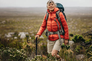 Abenteuerlustige ältere Frau auf einem Wanderausflug. Ältere Frau mit Jacke und Rucksack wandert auf dem Land und hält einen Wanderstock. - JLPSF15796