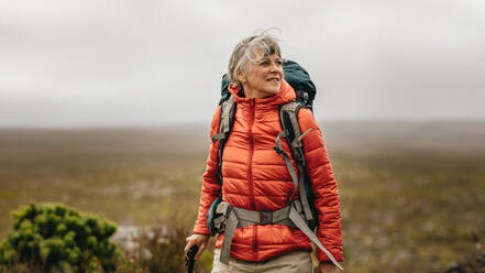 Frau, die auf einem Hügel spazieren geht und einen Trekkingstock hält. Frau mit Jacke und Rucksack auf einer Trekkingtour. - JLPSF15798