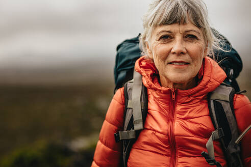 Porträt einer älteren Frau, die während eines Urlaubs im Freien steht. Ältere Frau mit Rucksack und Jacke, die während einer Trekkingtour steht. - JLPSF15800