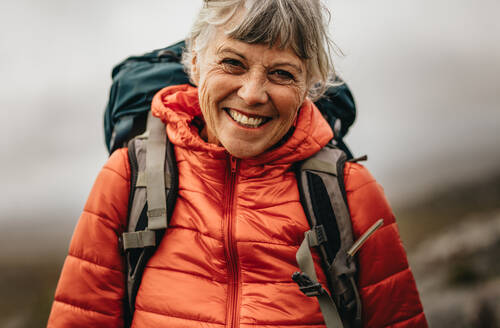 Close up of a senior woman wearing jacket and backpack standing outdoors. Smiling senior woman hiker standing outdoors with fog in the background. - JLPSF15802