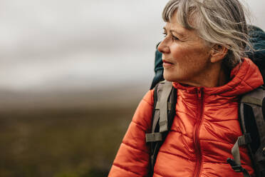 Nahaufnahme eines Porträts einer älteren Wanderin, die wegschaut. Frau mit Jacke und Rucksack auf einem Hügel an einem bewölkten Tag. - JLPSF15803