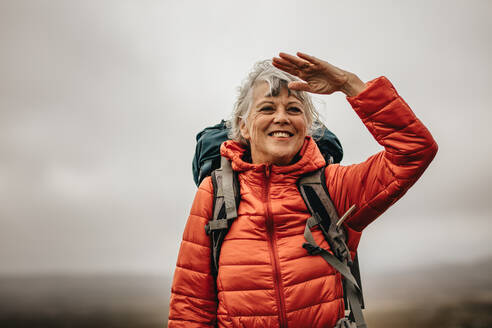 Lächelnde Touristin mit Rucksack, die an einem bewölkten Tag auf einem Hügel steht. Aufgeregte Wanderin, die auf einem Hügel stehend die Aussicht genießt. - JLPSF15805
