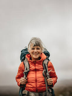 Lächelnde Wanderin mit Jacke und Rucksack auf einem Wanderausflug. Abenteuerlustige Frau auf einem Hügel an einem bewölkten Tag. - JLPSF15806