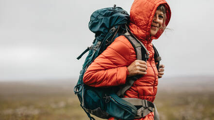 Side view of a woman hiker standing on top of a hill. Senior woman wearing a hooded jacket and a backpack standing on top of a hill during a trek. - JLPSF15810