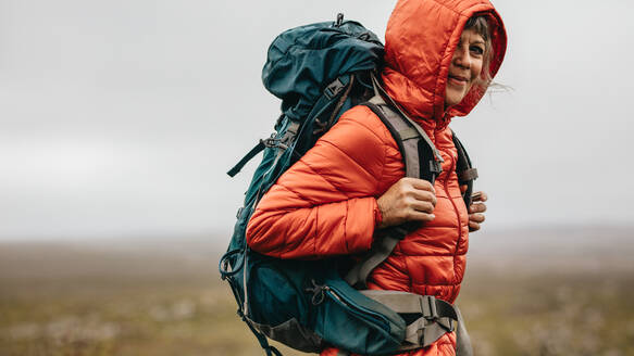 Seitenansicht einer Wanderin, die auf einer Anhöhe steht. Ältere Frau mit Kapuzenjacke und Rucksack, die während einer Wanderung auf einer Anhöhe steht. - JLPSF15810