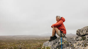 Seitenansicht einer Wanderin, die nach einer Wanderung auf einem Hügel sitzt. Lächelnde ältere Frau, die ihre Wanderung genießt und sich auf einem Hügel entspannt. - JLPSF15814