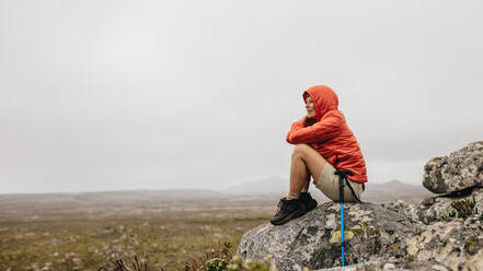 Frau sitzt auf einem Felsen auf einer Bergkuppe und betrachtet die Schönheit der Natur. Ältere Frau sitzt entspannt nach ihrer Wanderung mit einem Wanderstock an ihrer Seite. - JLPSF15815