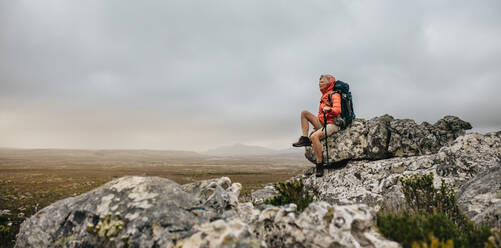 Eine ältere Frau sitzt auf einem Felsen und genießt die Aussicht von oben. Eine Wanderin entspannt sich, nachdem sie ihre Wanderung auf einem Hügel beendet hat. - JLPSF15817