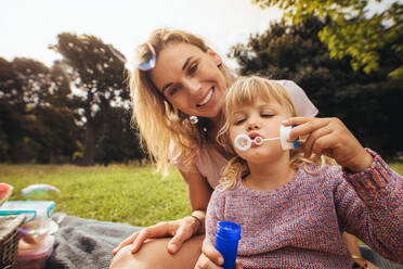 Cute little girl blowing soap bubbles while sitting with her mother outdoors. Mother and daughter having fun at picnic in park. - JLPSF15819