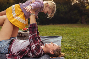 Junge Familie genießt einen Tag in der Natur, macht ein Picknick und verbringt Zeit miteinander. Mutter hebt ihre Tochter im Garten neben einem Picknickkorb hoch. - JLPSF15829