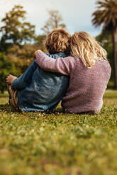 Rückansicht eines kleinen Jungen und eines Mädchens, die zusammen auf dem Rasen im Park sitzen. Kleiner Bruder und kleine Schwester sitzen im Park. - JLPSF15836