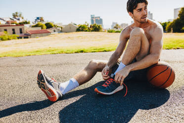 Athlete tying shoe laces sitting on ground with a basketball by his side. - JLPSF15837