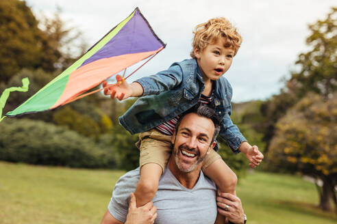 Little boy holding a kite sitting on his father's shoulders at the park. Happy father and son flying kite outdoors. - JLPSF15839