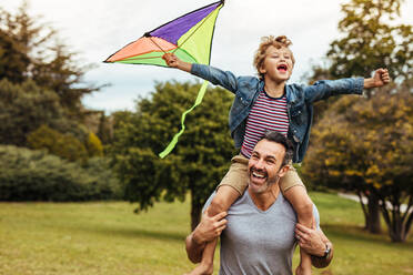 Young smiling boy playing with colorful kite outside sitting on father's shoulders. Little son with his father playing in a park with a kite. - JLPSF15840